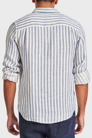 The rear view shows a person in an Academy Brand Farrelly Linen Shirt, featuring yarn-dye white and gray vertical stripes. The rolled-up sleeves and untucked style over dark pants create a relaxed look against a plain white background.