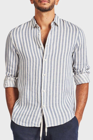 A person wears an Academy Brand Farrelly Linen Shirt with white and light blue vertical stripes. The long sleeves are rolled halfway up, paired with dark pants, hands in pockets against a plain white background.