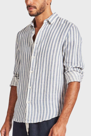 A man dons an Academy Brand Farrelly Linen Shirt in blue and white yarn dye stripes, sleeves rolled to the elbows. Half-buttoned, it complements dark pants on a plain white backdrop.