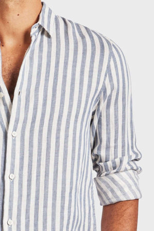 A person in a blue and white yarn-dye stripe Academy Brand Farrelly Linen Shirt with rolled-up sleeves and the top unbuttoned is partially visible against a plain white background.