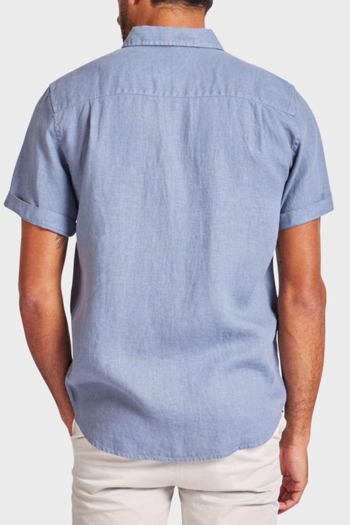A man is seen from the back wearing a light blue Academy Brand Hampton Linen Short Sleeve Shirt and white pants, with his hands in his pockets against a plain light background.
