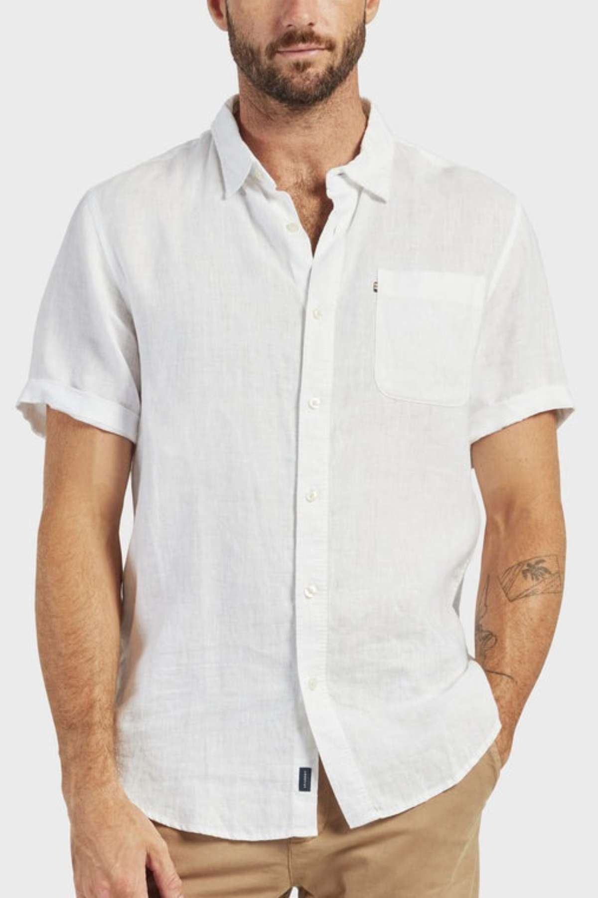 A man wears the Academy Brand Hampton Linen Short Sleeve Shirt—white, button-up with a chest pocket—and beige pants, standing against a light gray background. Tattoos are visible on his left arm.