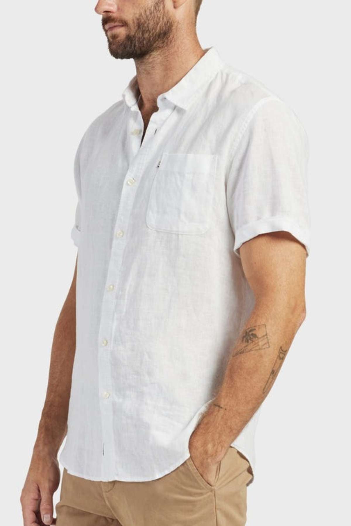 A man in an Academy Brand Hampton Linen Short Sleeve Shirt, white with rolled-up sleeves and a front pocket, teamed with light brown pants, stands one hand in his pocket, showing his beard and forearm tattoo.