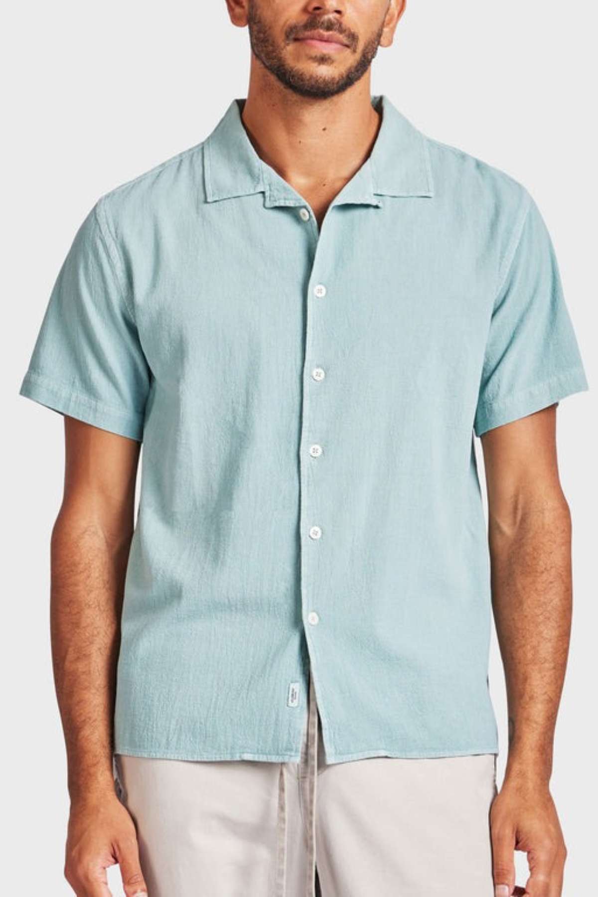 A man stands against a plain background wearing the Academy Brand Jed Short Sleeve Shirt, which features a revere collar and a relaxed fit in light blue. He pairs it with light-colored pants for a casual look.