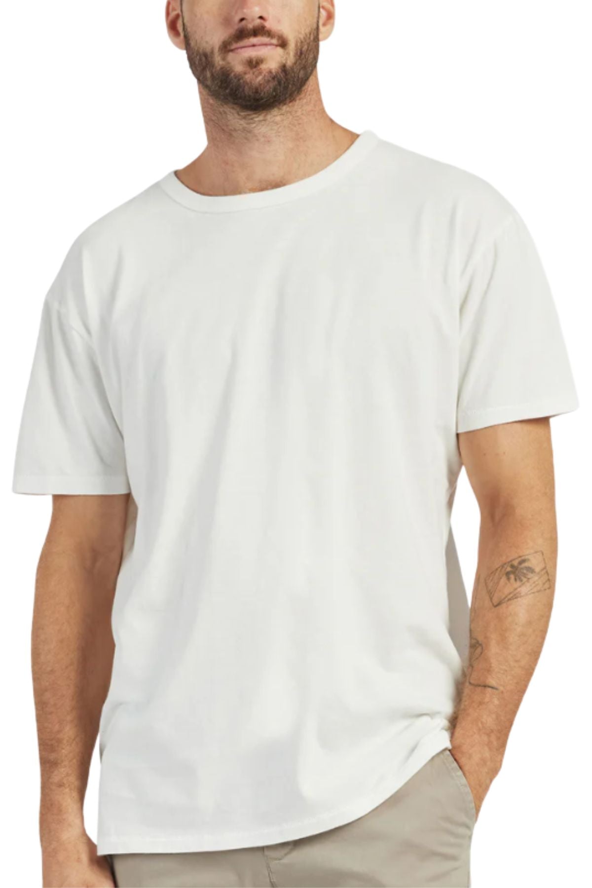 A man with a short beard and a visible left forearm tattoo stands with one hand in his pocket, dressed in light pants and a plain white Academy Brand Jimmy Tee made from 100% cotton.