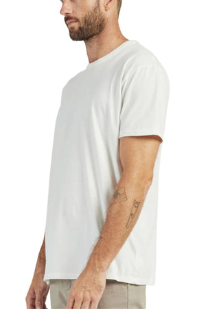 A bearded man stands in profile against a white background, wearing the Academy Brand Jimmy Tee—a loose fit, 100% cotton shirt—with light-colored pants and tattoos visible on his right forearm.