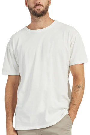 A man with a short beard and a visible left forearm tattoo stands with one hand in his pocket, dressed in light pants and a plain white Academy Brand Jimmy Tee made from 100% cotton.