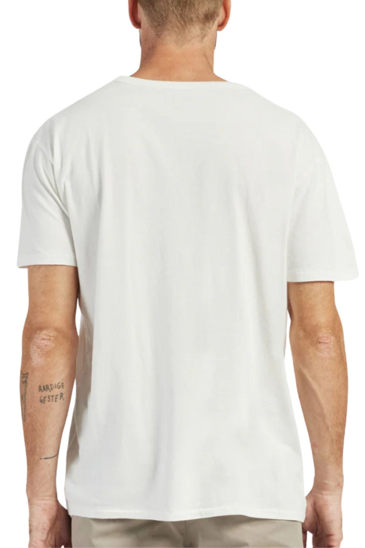 A man faces away from the camera, wearing the Academy Brand Jimmy Tee—100% cotton with a loose fit. He has a small triceps tattoo and pairs the white tee with light-colored pants.
