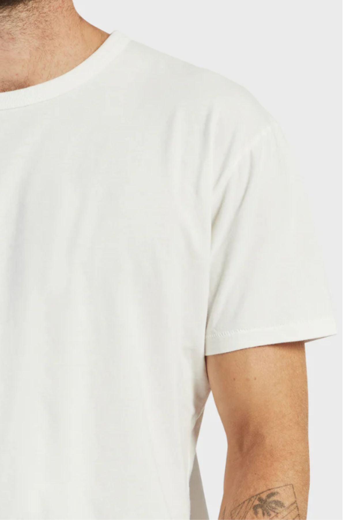 A person wearing an Academy Brand Jimmy Tee, a loose 100% cotton white shirt; only their upper torso and part of the left arm with a small forearm tattoo are visible.