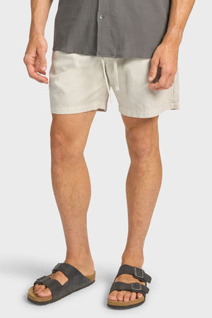 Wearing Academy Brand Oahu Short in a relaxed volley fit, paired with a dark gray shirt and black cork-soled sandals, a person stands against a plain light background. Their head is not visible in the image.