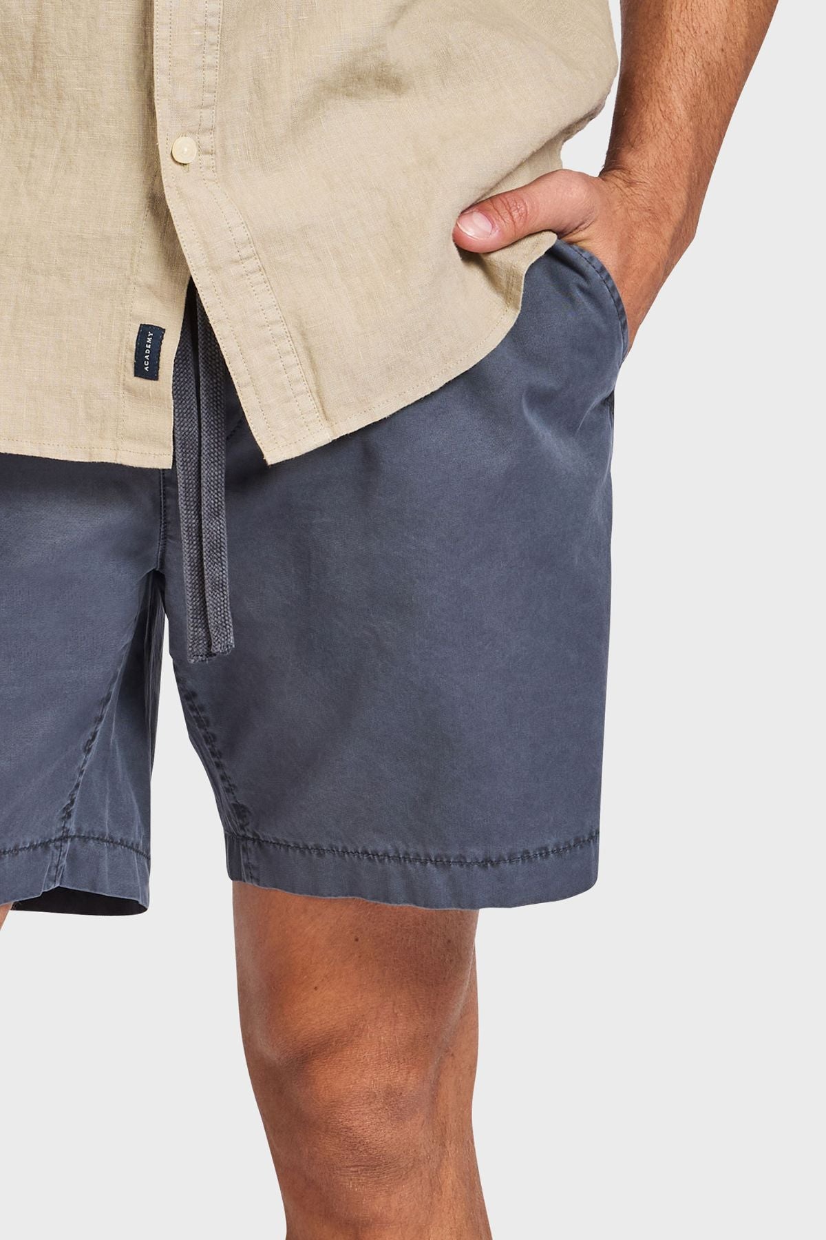 A person wears a beige button-up shirt and Academy Brand Oahu Short cotton twill shorts, standing with one hand in their pocket against a plain light background.