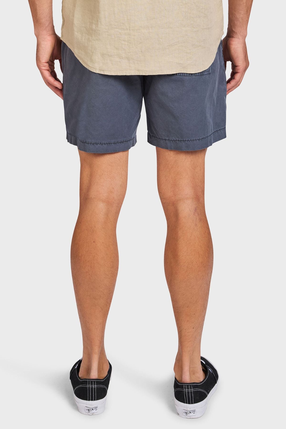 A person stands with their back to the camera, wearing a beige shirt and gray Academy Brand Oahu Short, paired with black sneakers with white soles. The plain white background highlights the outfit's vintage style.