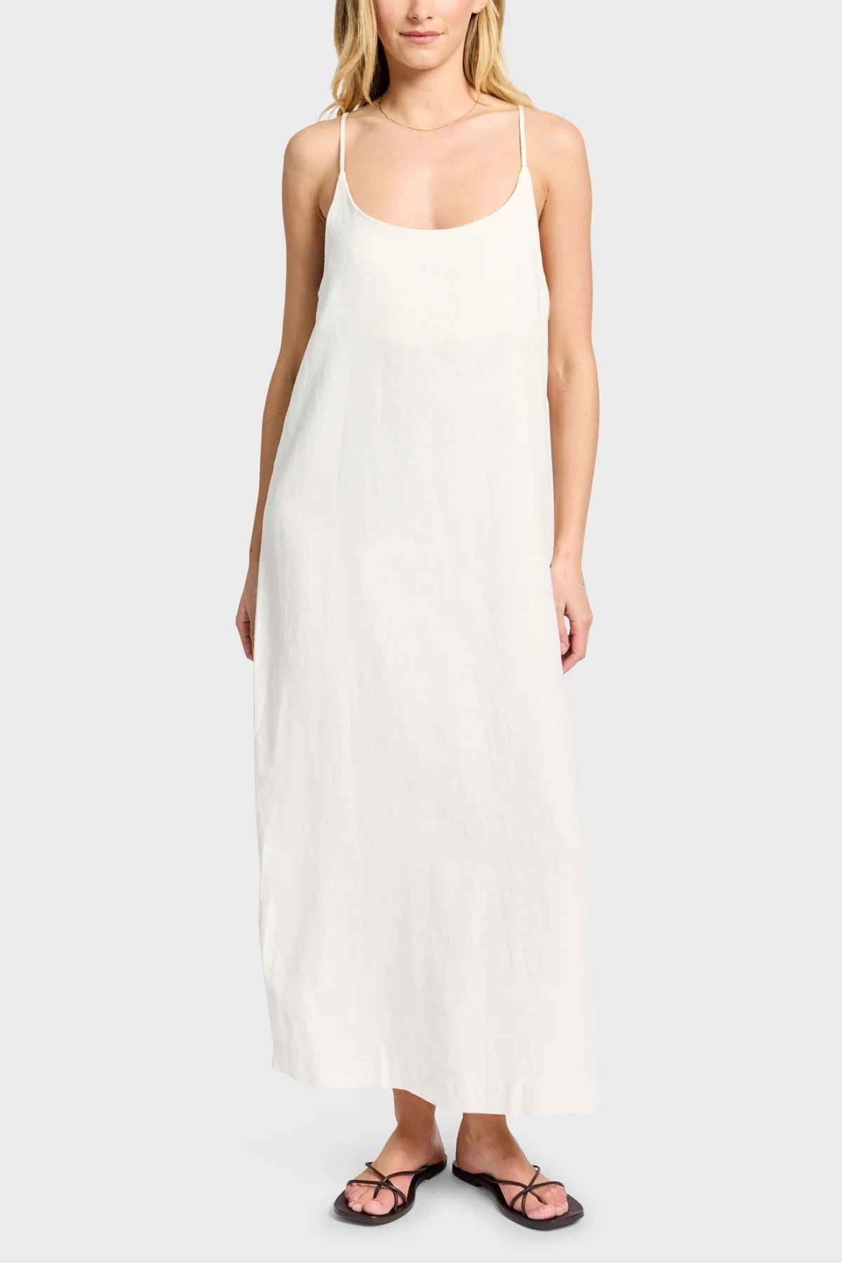 A person stands against a plain background wearing the Academy Women Essential Linen Slip Dress, a long white linen garment with adjustable straps and subtle side splits. The outfit is completed with black flip-flops.