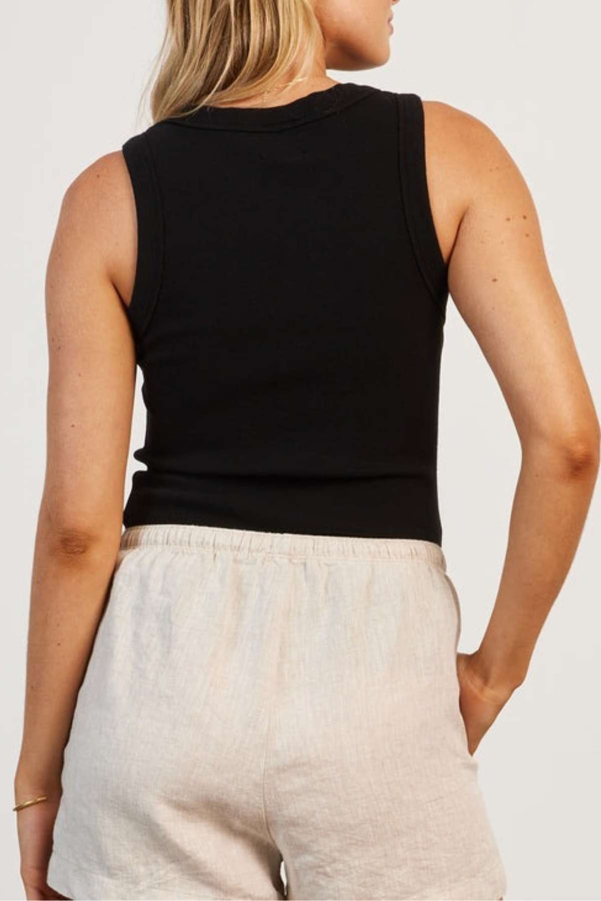 A person with light hair is seen from behind wearing the Academy Women Essential Rib Tank and light beige shorts. The premium garment-washed rib tank complements their casual summer wardrobe as they stand with one hand resting on their hip against a plain background.