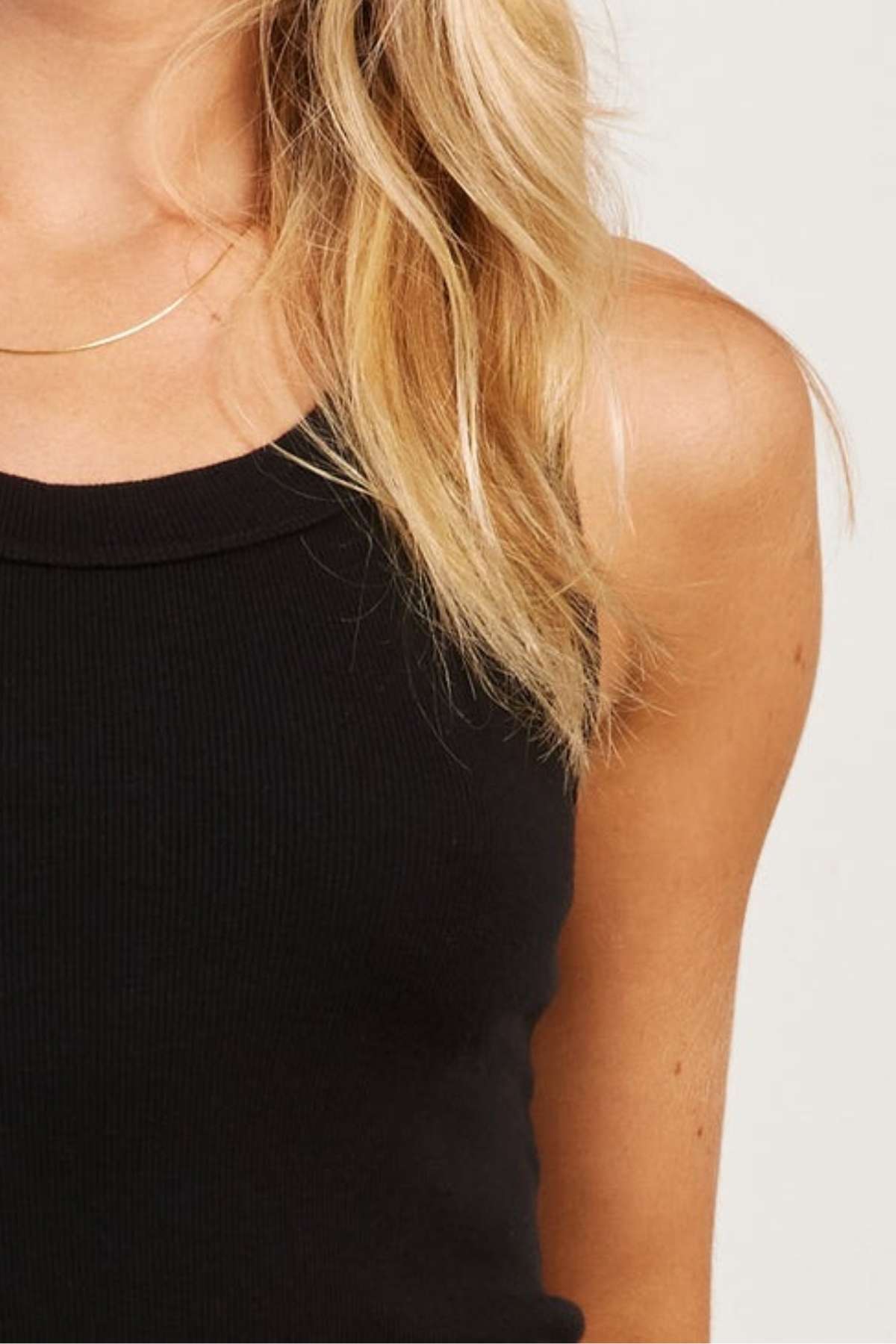 Close-up of an individual with long blond hair donning an Academy Women Essential Rib Tank. The image captures the left shoulder, a section of the chest, and flowing hair over the shoulder. This premium garment-washed rib top is an ideal addition to your summer wardrobe. The background features a light neutral hue.