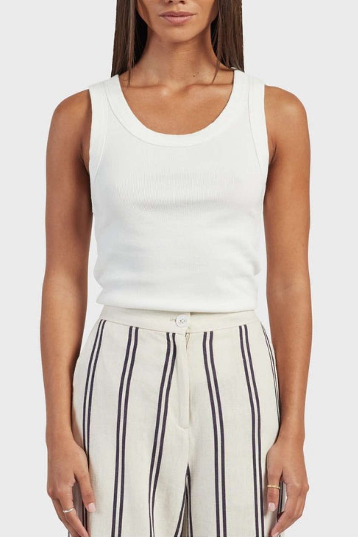 A person wearing an Academy Women Essential Rib Tank, which is a white sleeveless premium garment washed rib top, and high-waisted cream-colored pants featuring vertical navy and white stripes. They have long straight hair with their hands relaxed at their sides, embodying the ideal summer outfit. The background is a plain light gray.
