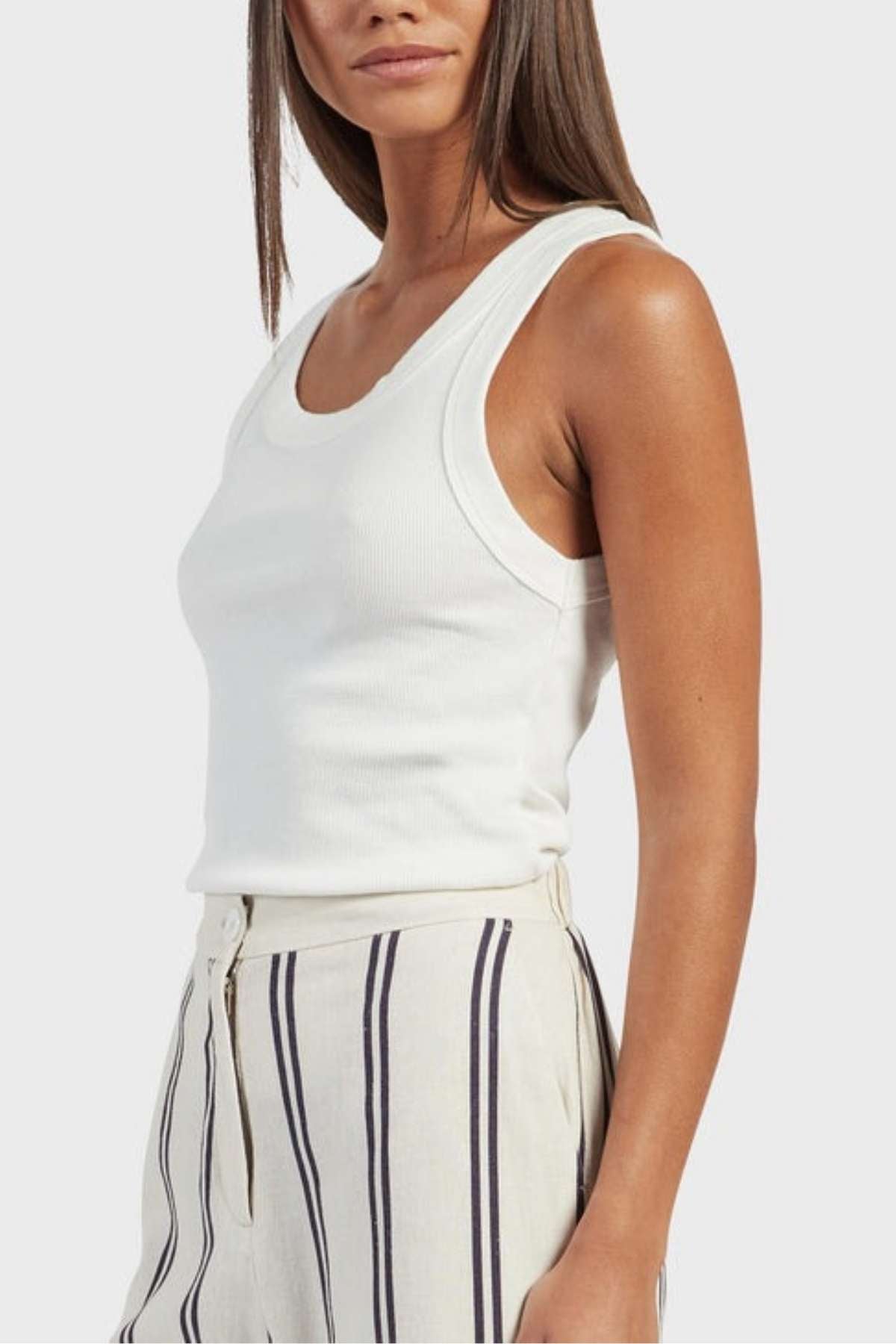 A woman is wearing the Academy Women Essential Rib Tank, matched perfectly with high-waisted, light-colored striped pants. Her long, straight hair cascades down as she poses slightly turned to the side, highlighting her casual yet stylish summer ensemble.