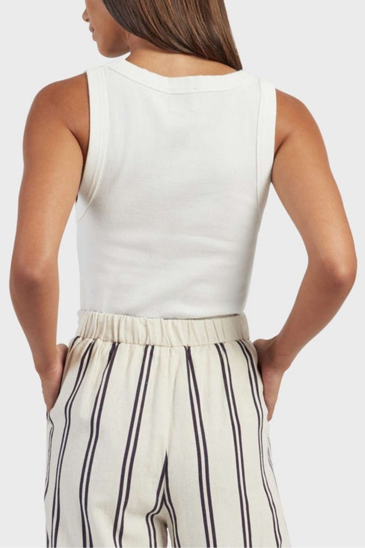 Rear view of a person with long brown hair, wearing an Academy Women Essential Rib Tank paired with high-waisted, cream-colored pants adorned with vertical black and white stripes - a flawless addition to any summer wardrobe.