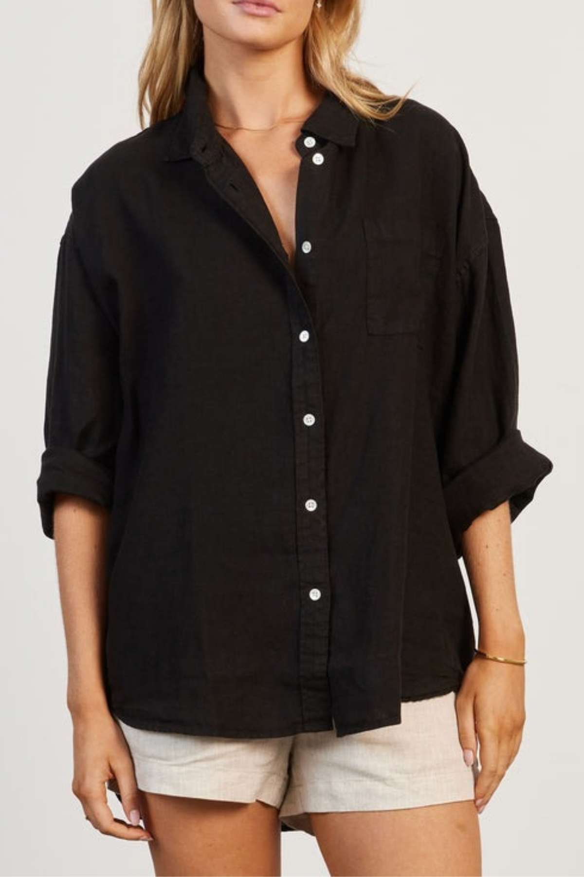 A person stands wearing a black, loose-fitting Academy Women Hampton Linen Shirt adorned with mother-of-pearl buttons and rolled-up sleeves. The shirt, which is garment dyed for a distinctive look and includes a pocket on the left chest, is paired with beige shorts. They pose against a plain background in a relatively relaxed manner.