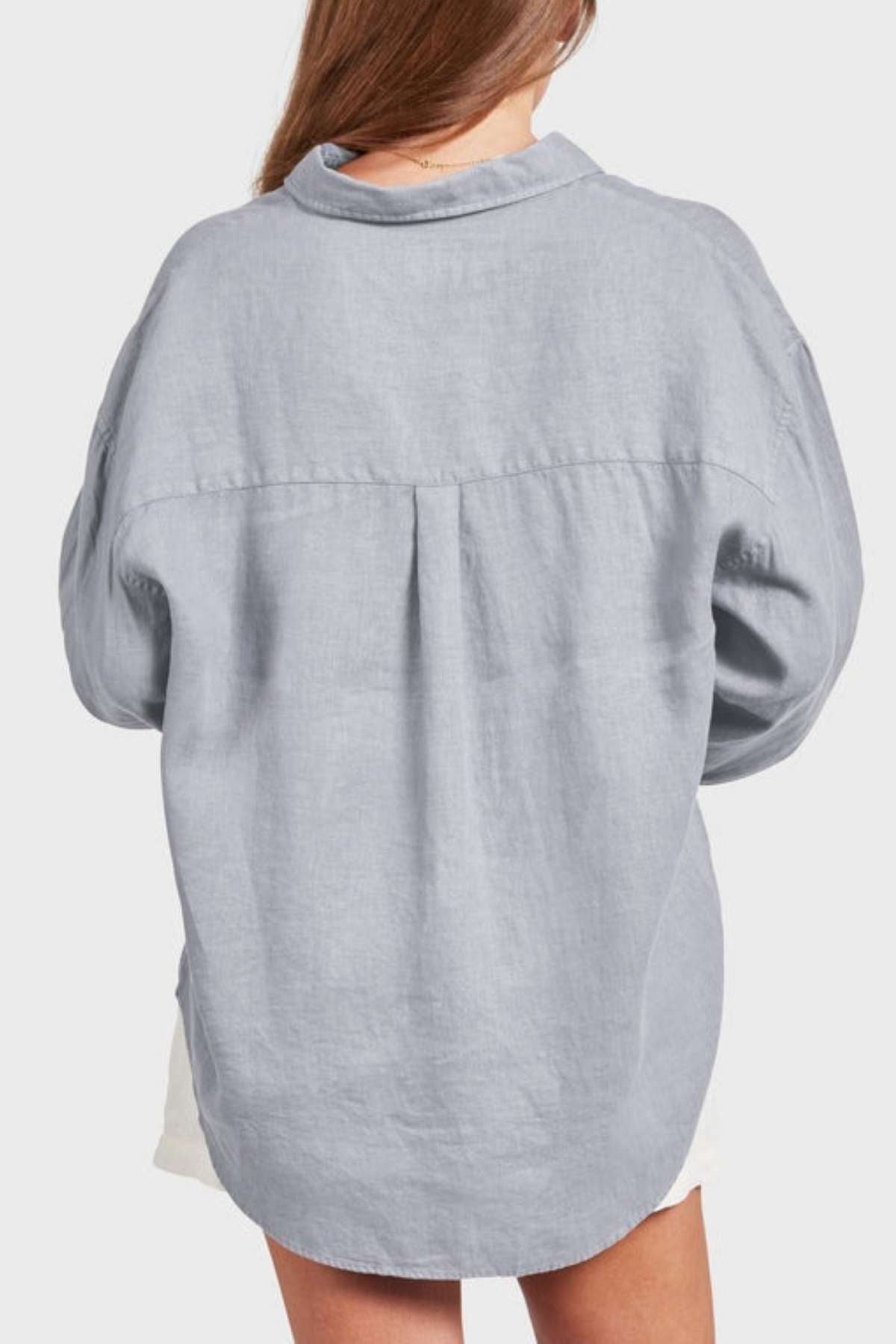 Seen from behind, a person with long hair wears a loose, gray Academy Women Hampton Linen Shirt. The garment-dyed button-up shirt features a collar and dropped shoulders and is paired with white shorts. Set against a plain, light-colored background, this shirt is known for its extra softness.