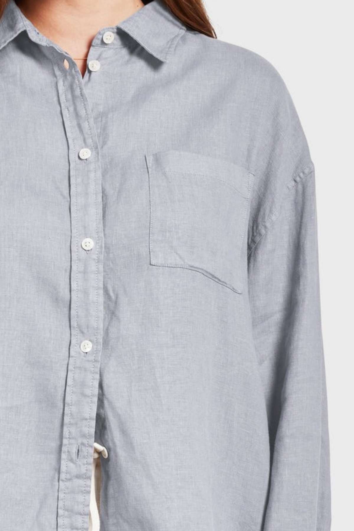A person wearing a light blue garment-dyed Academy Women Hampton Linen Shirt with a collar and single chest pocket is partially visible. The focus is on the upper part of this shirt, highlighting its buttons and pocket. The background is plain and light-colored, enhancing the extra softness of the fabric.