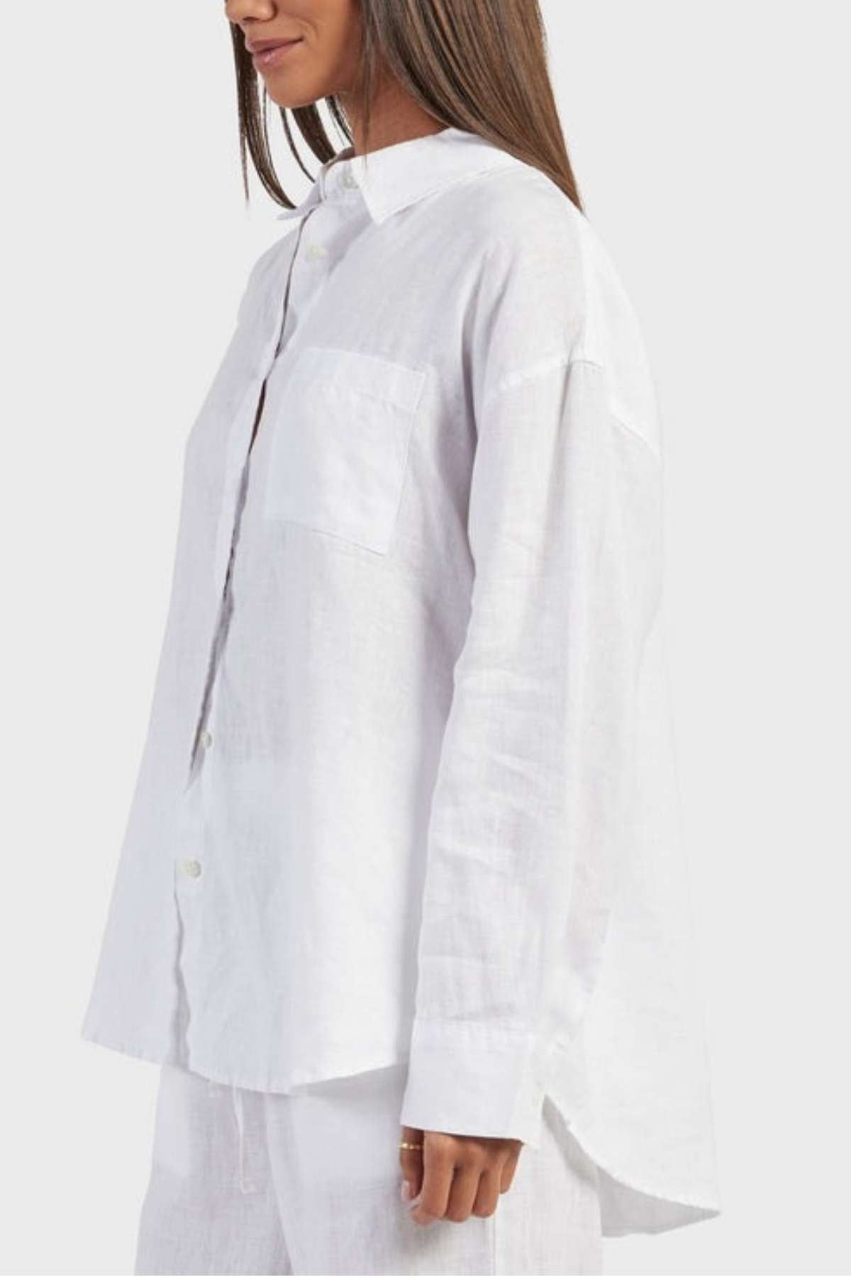 A person with long, straight hair is wearing a loose-fitting, white Academy Women Hampton Linen Shirt and matching white pants. The shirt, featuring natural mother-of-pearl buttons and a chest pocket, is slightly wrinkled. The photo is taken from a side angle against a plain, light background.