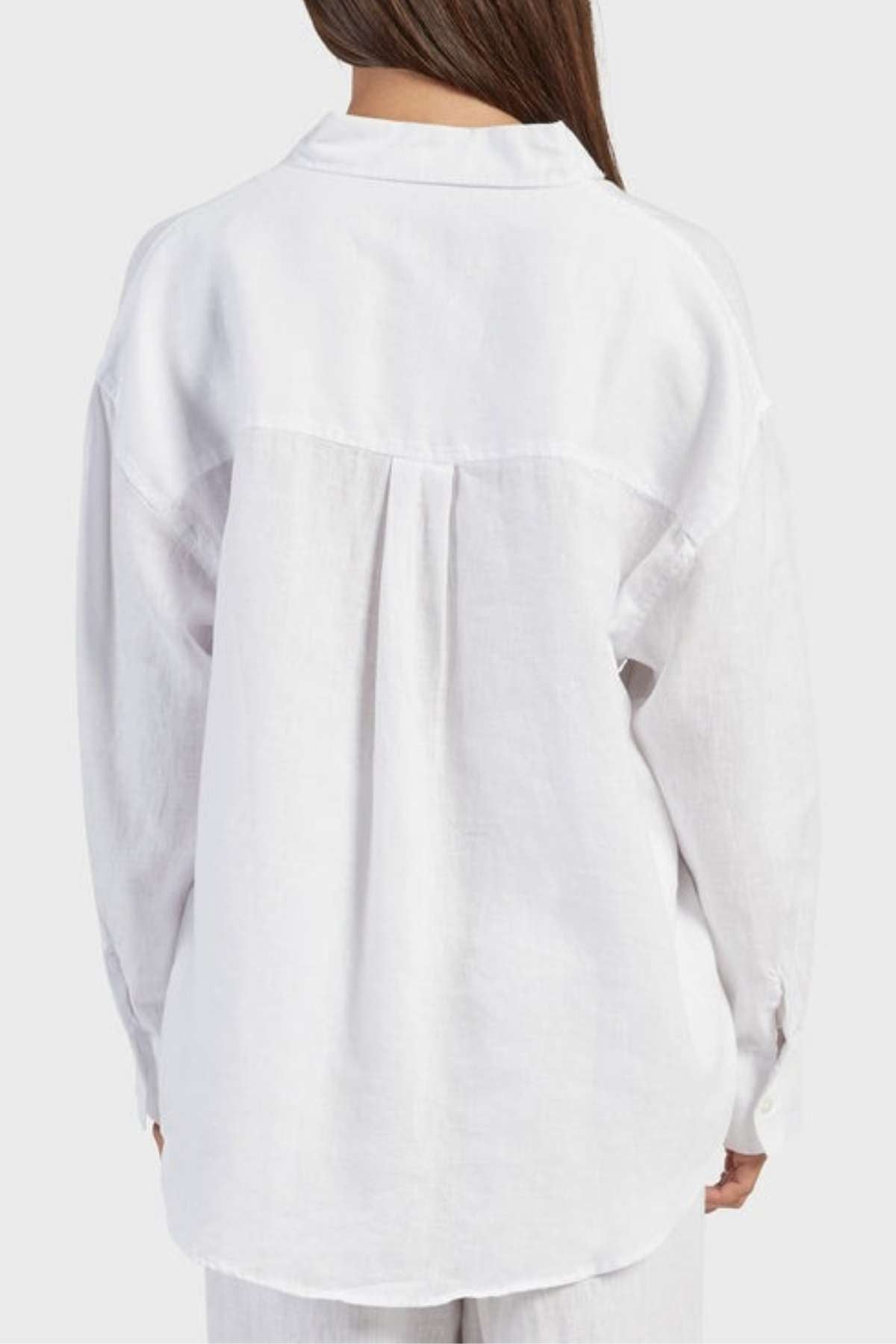 A person with long brown hair is standing with their back to the camera, wearing a loose-fitting, white Academy Women Hampton Linen Shirt. The shirt features natural mother-of-pearl buttons and a gathered detail at the yoke, providing a relaxed, casual fit. The background is plain and light gray.