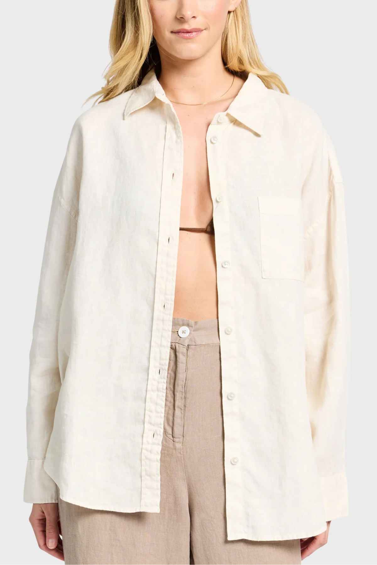 A person wearing the Academy Women Hampton Linen Shirt, featuring mother-of-pearl buttons, open to reveal a brown bikini top underneath, paired with beige pants. The plain gray background highlights the timeless elegance of this garment-dyed ensemble.