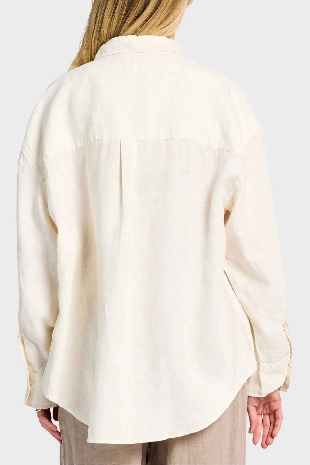 A person with long hair is seen from behind wearing a cream-colored Academy Women Hampton Linen Shirt, which features mother-of-pearl buttons. The shirt offers a loose fit and pairs perfectly with beige pants against a plain white background.