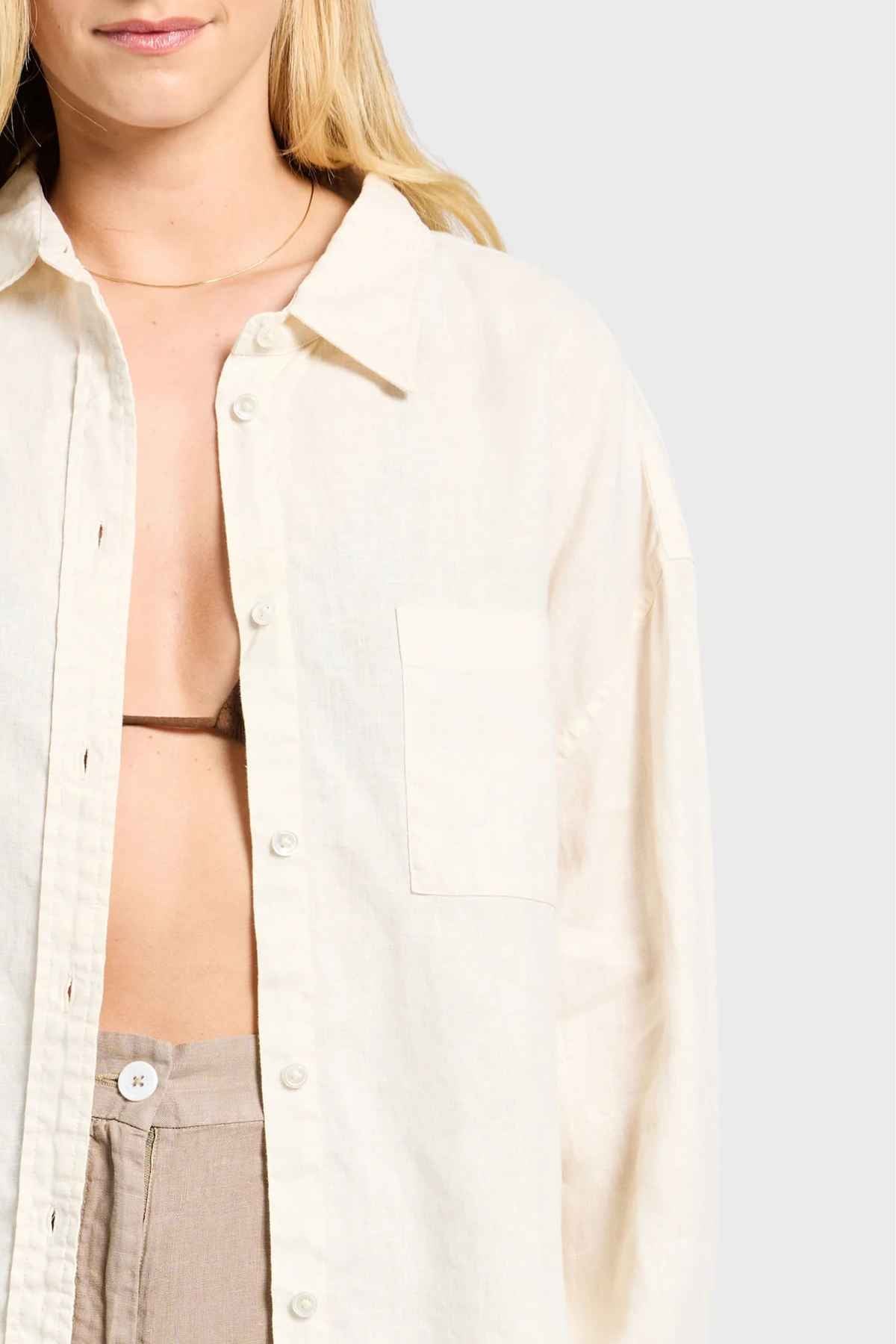 A person with long blonde hair wears an Academy Women Hampton Linen Shirt, its mother-of-pearl buttons undone to reveal a brown bikini top underneath. The background is plain light gray.