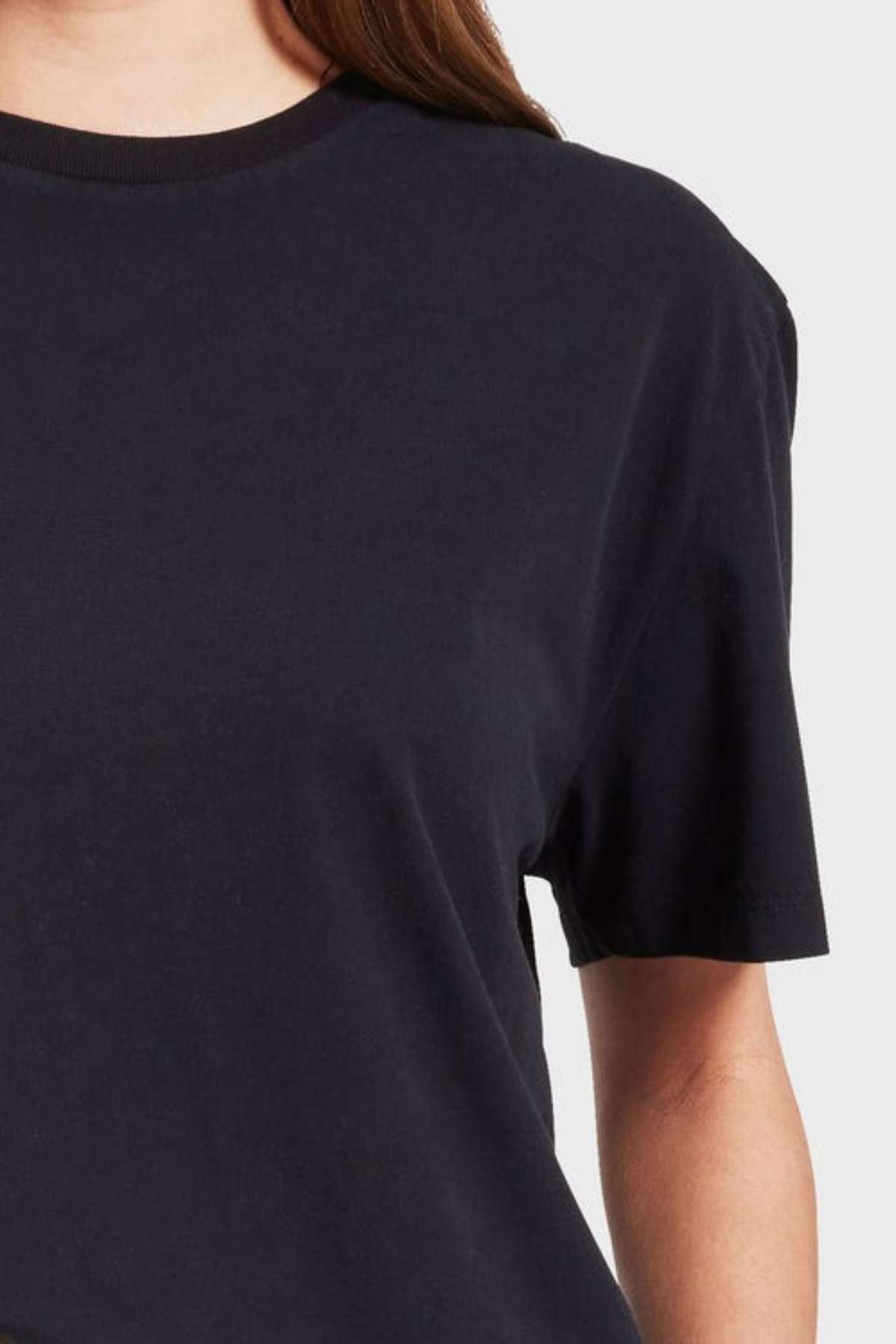 Close-up of a person wearing a plain black Academy Women Jimmy Boyfriend Tee, showing the shoulder and part of the arm against a plain, light background. The premium sanded jersey T-shirt has a casual, relaxed fit, and the person's hair is partially visible.