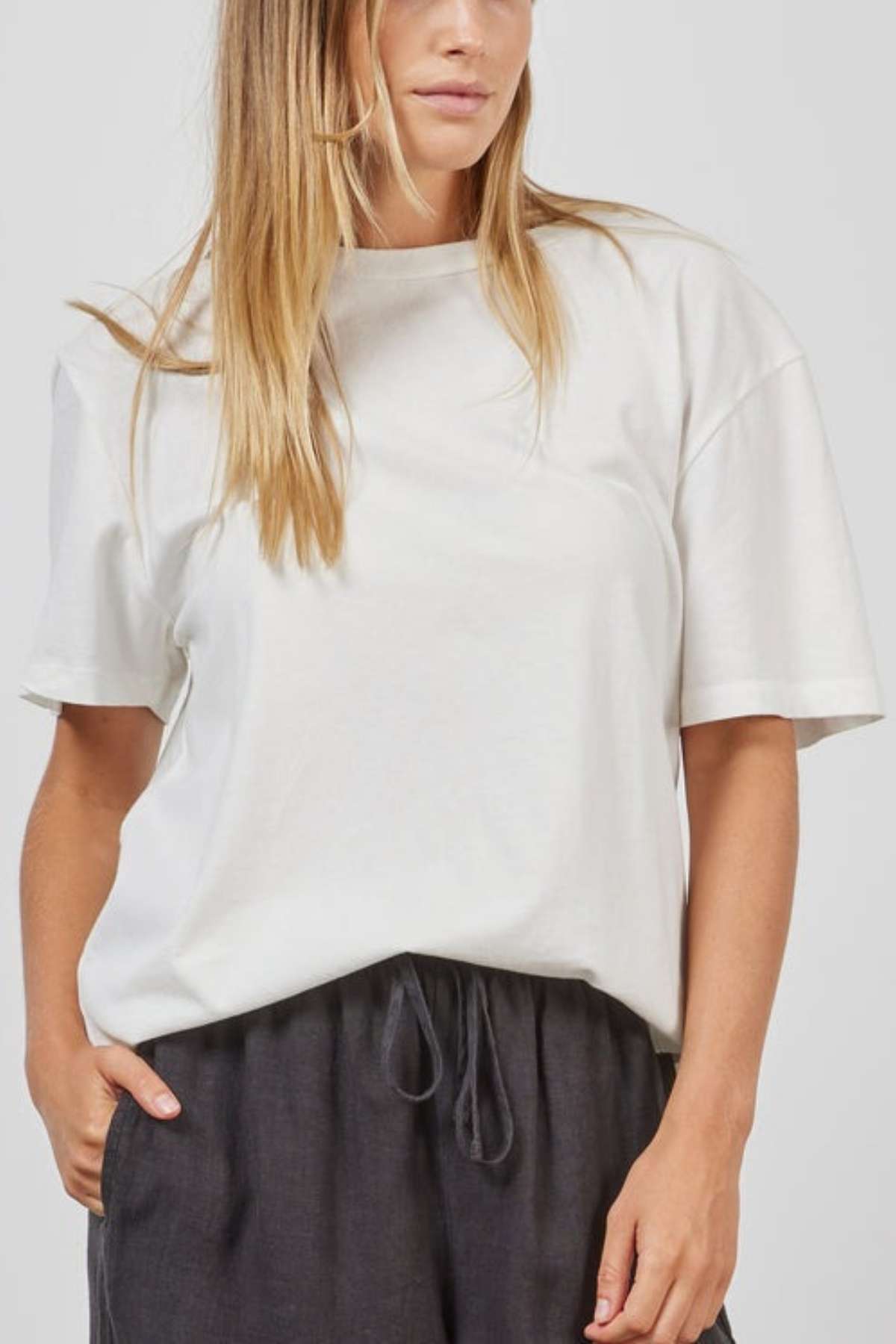 A woman with long blonde hair wears the Academy Women Jimmy Boyfriend Tee in a premium sanded jersey fabric, paired with dark gray drawstring waistband pants. Her hands are in her pockets, and she is standing against a plain background.