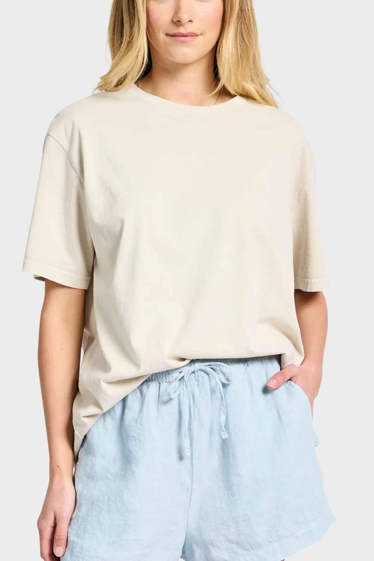 A person with long hair is wearing an Academy Women Jimmy Boyfriend Tee in premium sanded jersey, paired with light blue shorts featuring a drawstring, standing against a plain light gray background. One hand casually rests in the shorts pocket.