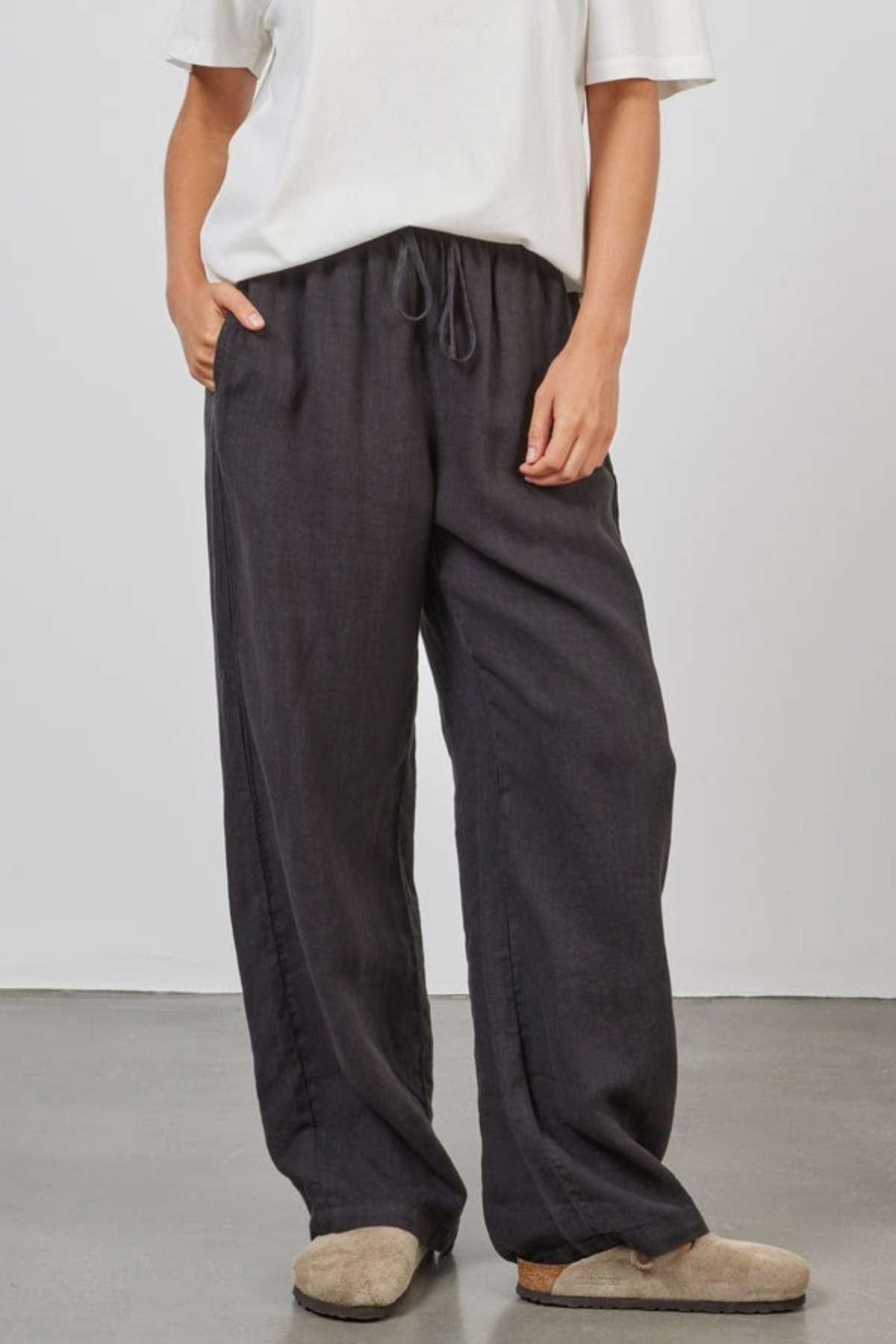 A person is wearing a white short-sleeve shirt and a pair of Academy Women Riviera Linen Pants, which are relaxed wide-leg black pants with a drawstring waist. They have one hand in their pocket and are standing on a plain, smooth surface. The person is also wearing light brown slippers.