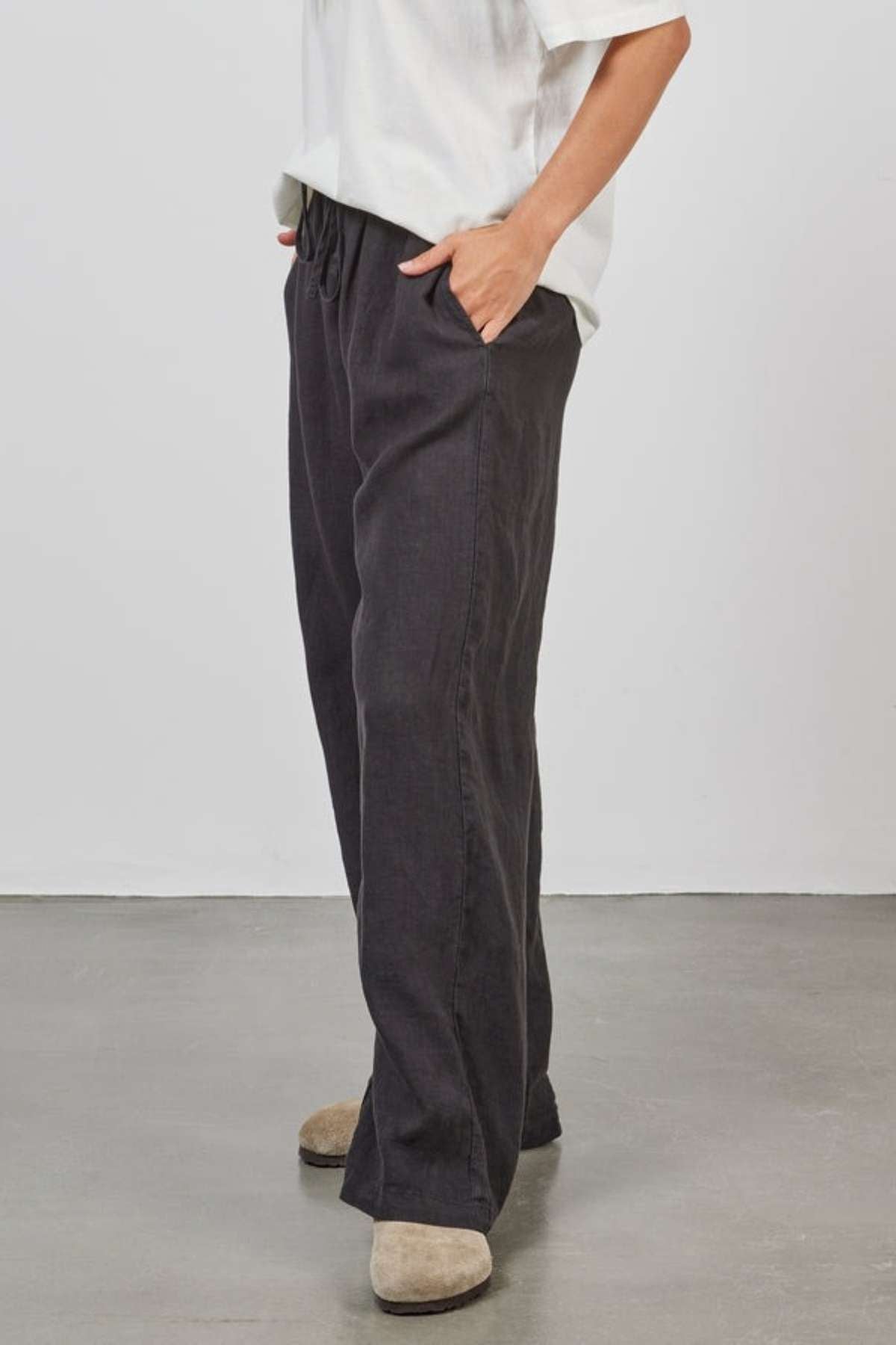 A person wearing a white T-shirt and the Academy Women Riviera Linen Pant in dark hue with hands in pockets stands on a polished concrete floor. Beige shoes are visible. The background is a simple white wall.