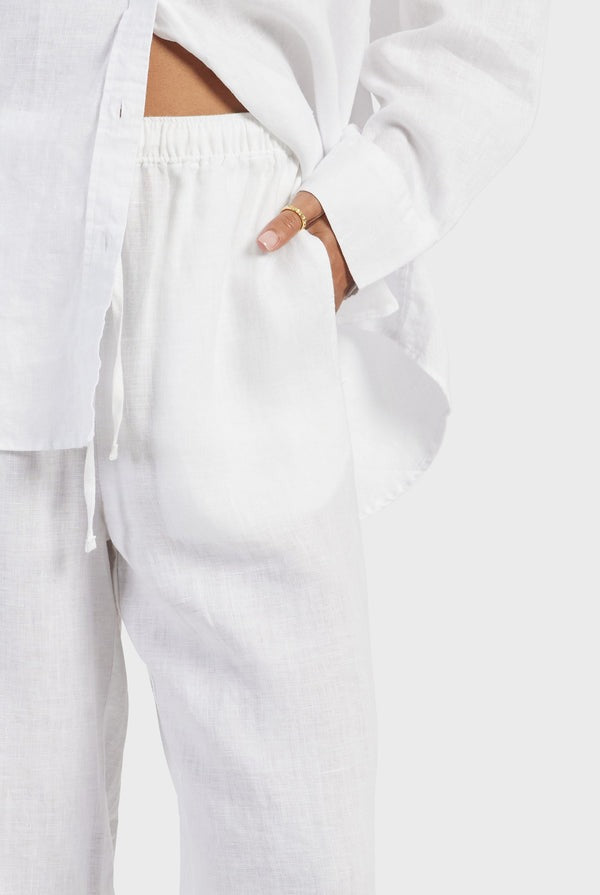 A person wearing a white long-sleeve button-up shirt and Academy Women Riviera Linen Pants stands with one hand casually in their pocket. The lightweight and breathable outfit appears comfortable and relaxed against a plain background.