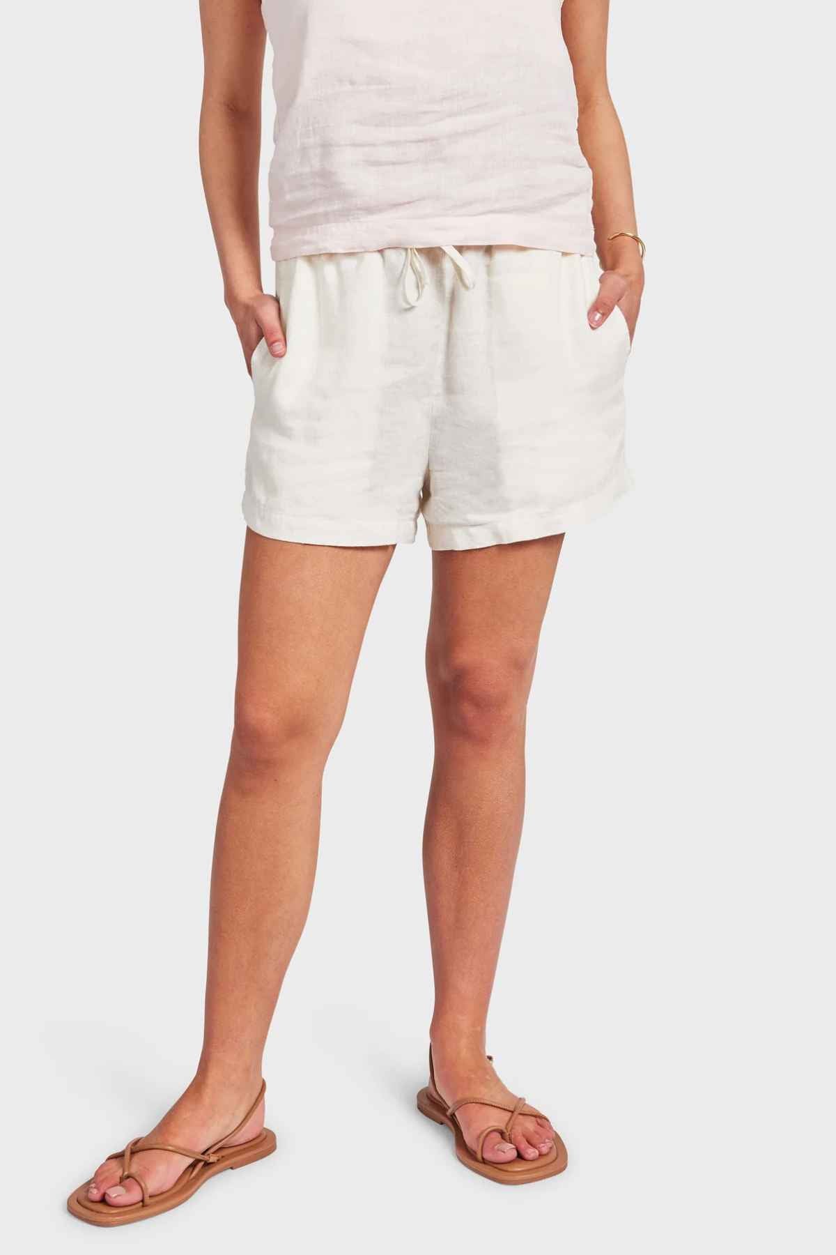 A person wears a sleeveless top and the Academy Women Riviera Linen Shorts, featuring an elastic waist and casual side entry pockets, standing against a plain background. They complete their relaxed look with a pair of comfortable sandals.
