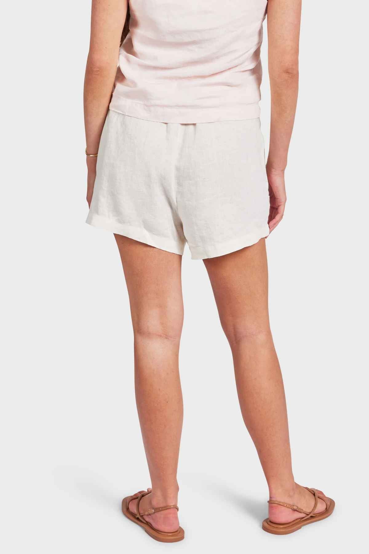 Rear view of a person wearing a light pink sleeveless top, Academy Women Riviera Linen Shorts with an elastic waist, and brown sandals. They stand with their arms relaxed by their sides against a plain white background.