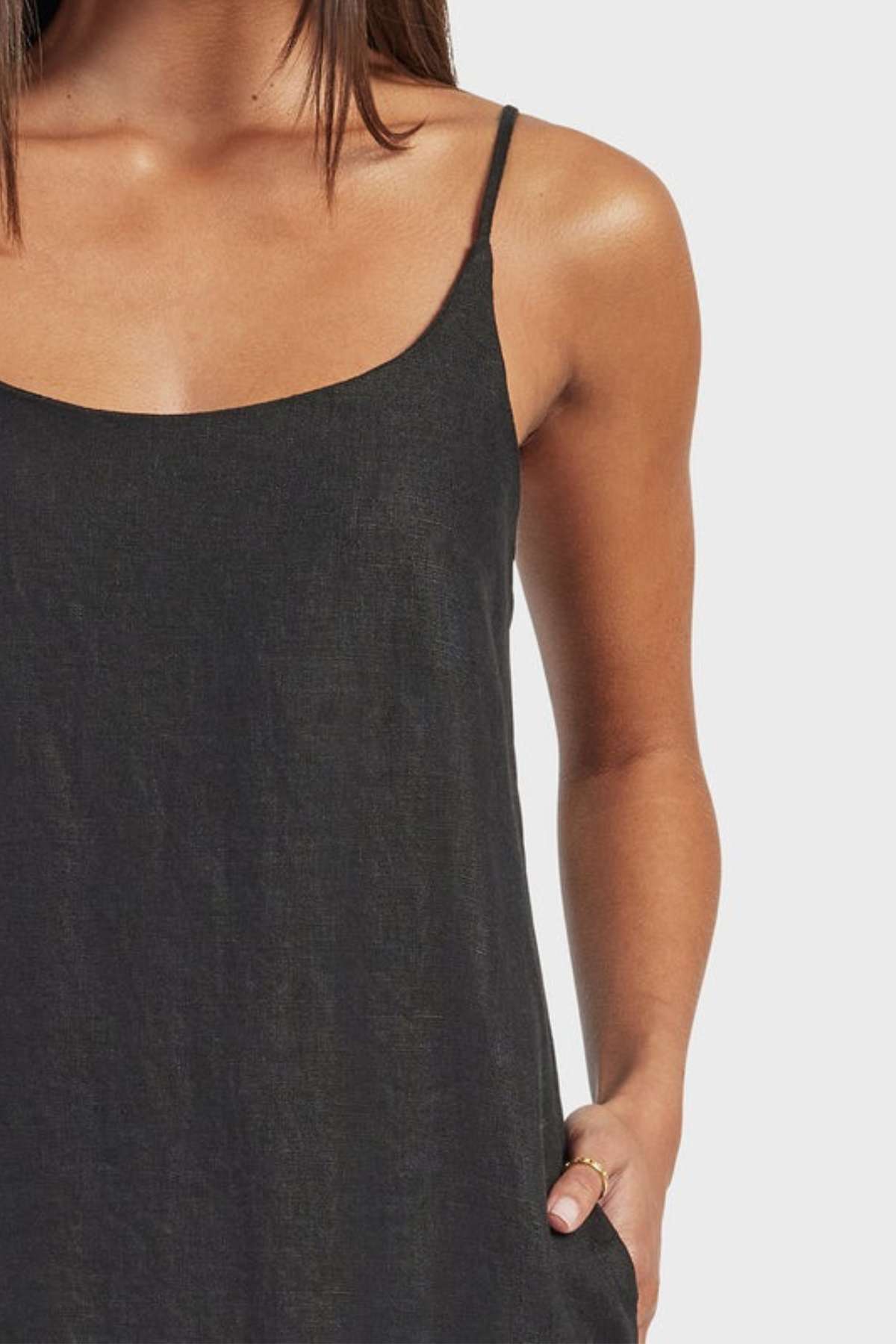 A woman wearing the Academy Women Essential Linen Slip Dress—sleeveless, black linen with adjustable straps and subtle side splits—stands against a light gray background, one hand in her pocket and her hair down.
