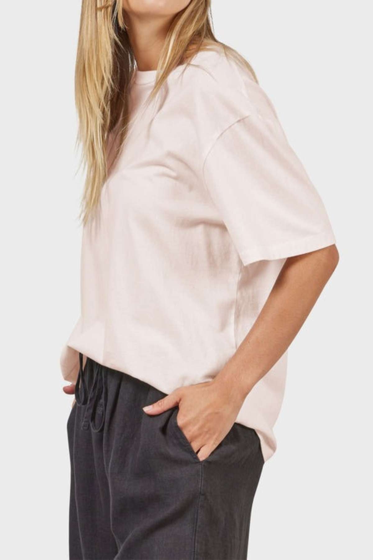 A person with long hair wears an oversized, light pink Academy Women Jimmy Boyfriend Tee crafted from premium sanded jersey and pairs it with dark pants. Their right hand rests in the pocket of their pants as they stand against a plain, light backdrop.