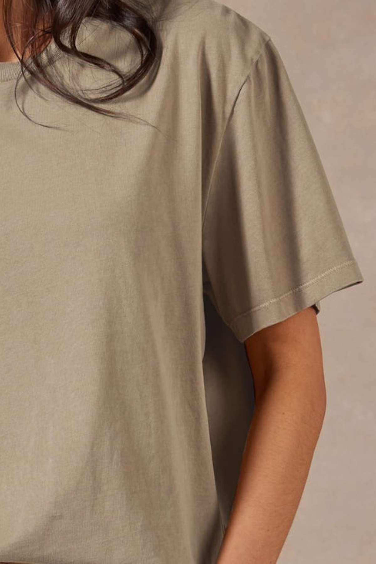 A person with long, dark hair wears the Academy Women Jimmy Boyfriend Tee—a loose, short-sleeved beige top made from premium sanded jersey—shown from shoulders to waist against a neutral background.