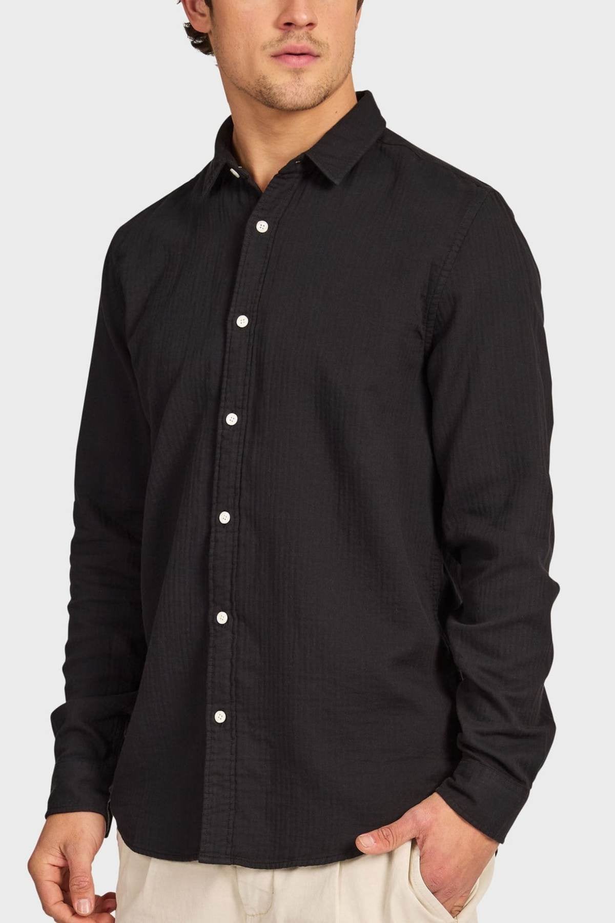 A man wearing a long-sleeved black Academy Brand Harry Double Cloth Shirt with white buttons and light pants stands with one hand in his pocket against a plain background.