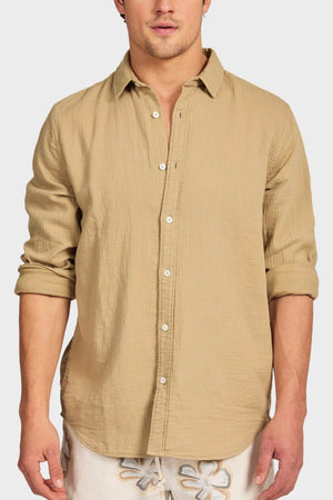 A man wearing the Academy Brand Harry Double Cloth Shirt in light brown with rolled-up sleeves, paired with light floral shorts, stands against a plain background.