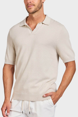 A man wearing the Academy Brand Jonny Textured Knit Polo in light beige with a relaxed collar and white pants stands with one hand in his pocket against a plain background.
