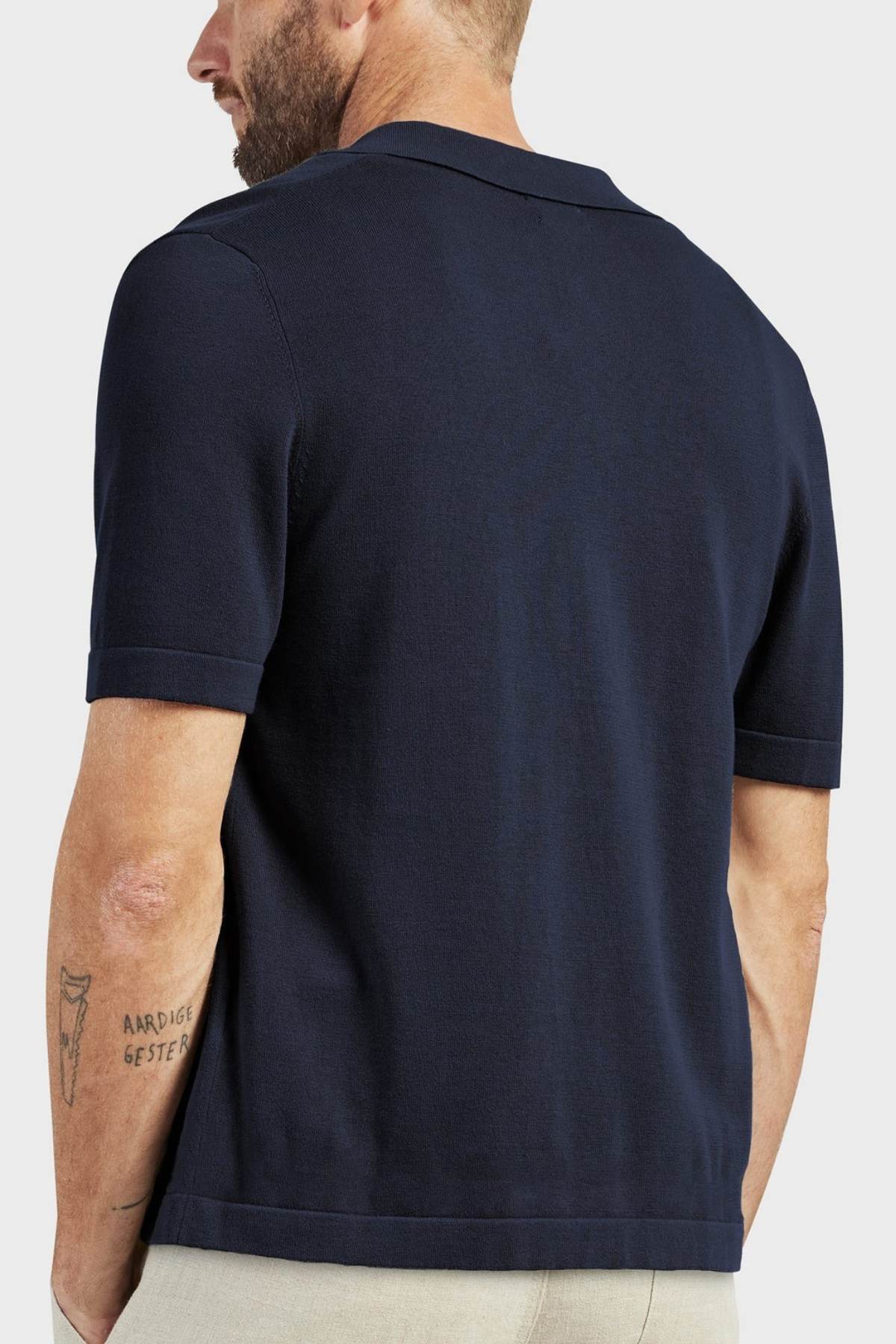 A man with a trimmed beard is seen from behind, wearing an Academy Brand Jackson Knit Short Sleeve Shirt in dark navy and light pants. He has a text tattoo on his left forearm, set against a plain, light background.