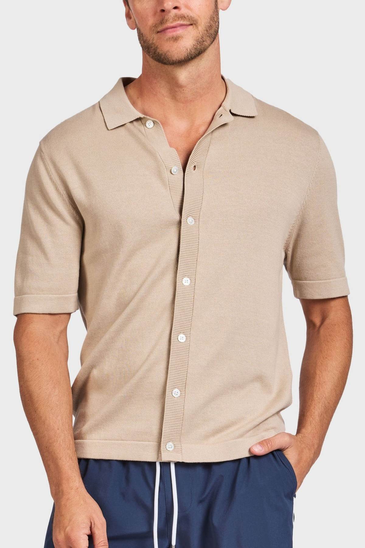A man wears the Academy Brand Jackson Knit Short Sleeve Shirt in beige with a collar and navy blue drawstring pants, standing with one hand in his pocket against a plain white background.