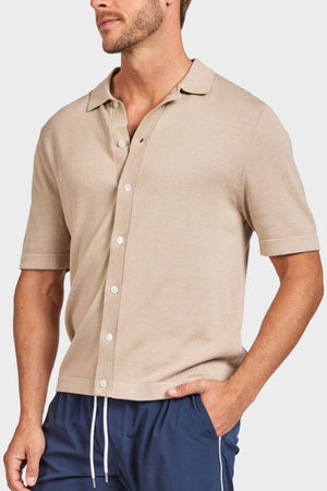 A man wears the Academy Brand Jackson Knit Short Sleeve Shirt in beige with navy drawstring pants, one hand in his pocket, facing slightly sideways against a plain background.
