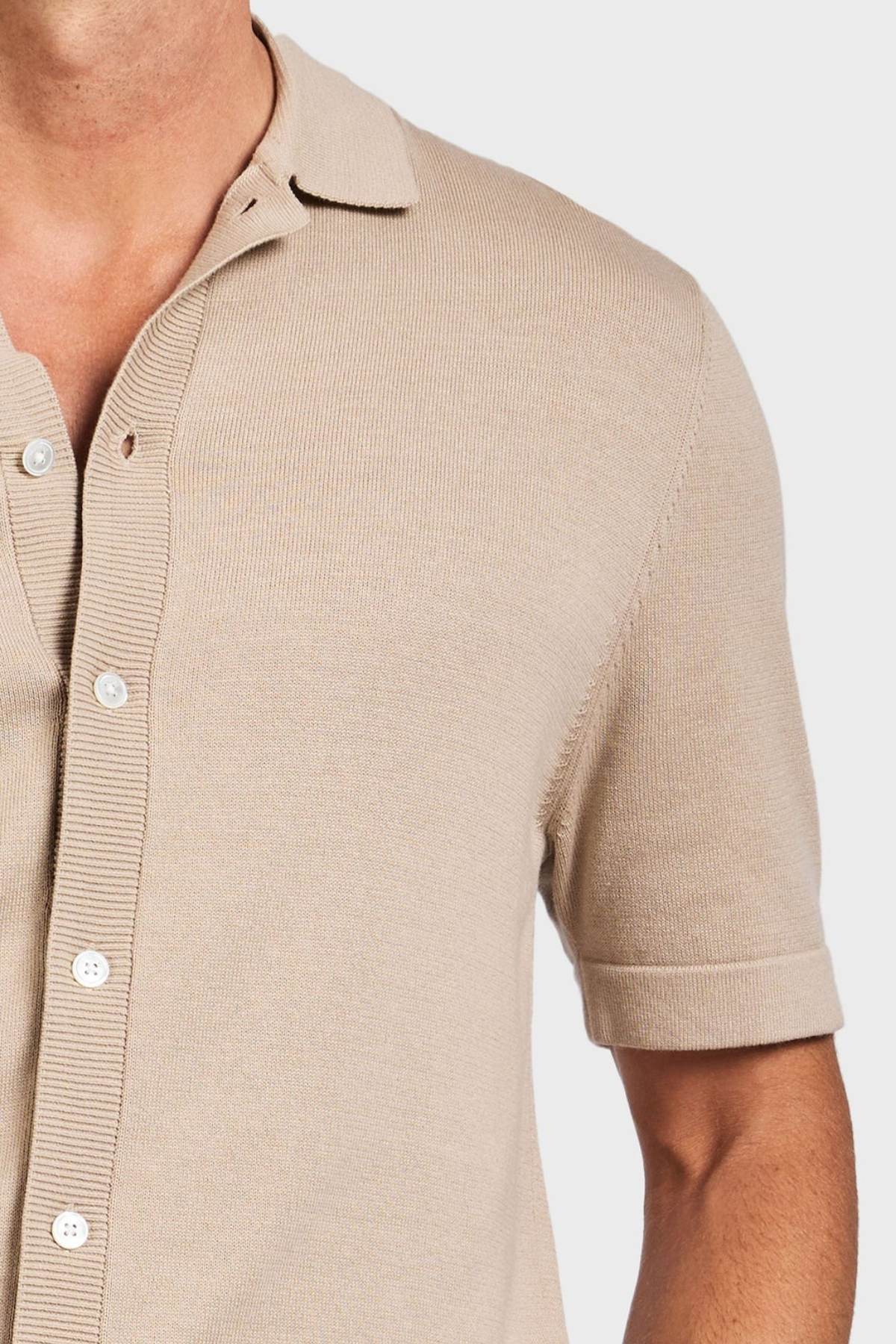 A close-up shows a person wearing the Academy Brand Jackson Knit Short Sleeve Shirt in beige with white buttons, highlighting the upper torso and part of the arm against a plain light background.
