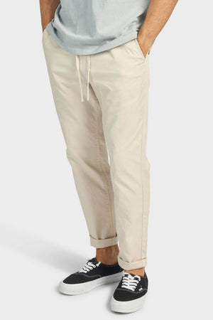 A person stands against a plain white background, hands in pockets, wearing the Academy Brand Oahu Pant in light beige soft cotton twill with a light grey t-shirt and black-and-white sneakers.