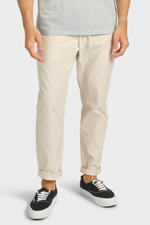 A person wearing Academy Brand Oahu Pant in cream cotton twill with a drawstring waist, paired with a light gray shirt and black-and-white sneakers, stands against a plain white background.